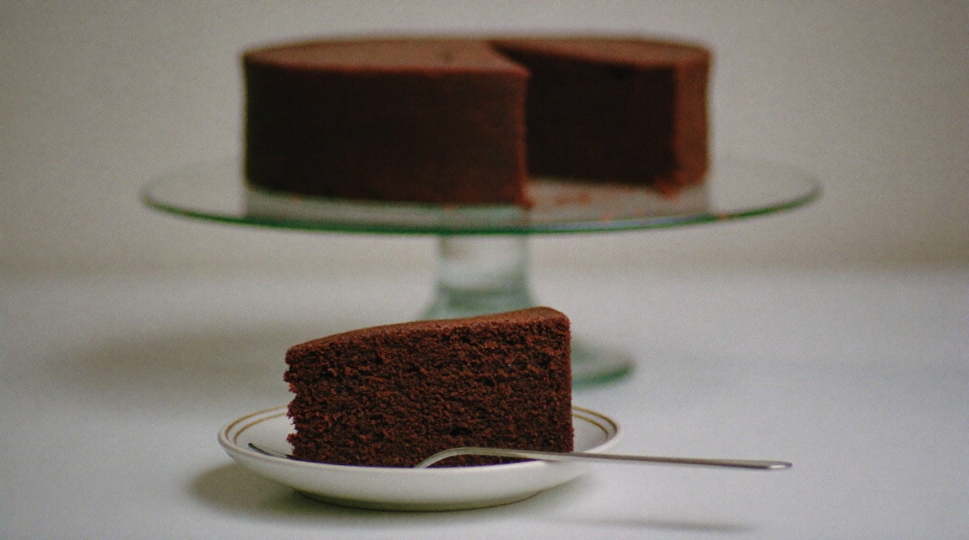 A single slice of layer cake on a side plate with the whole cake softly out of focus behind it